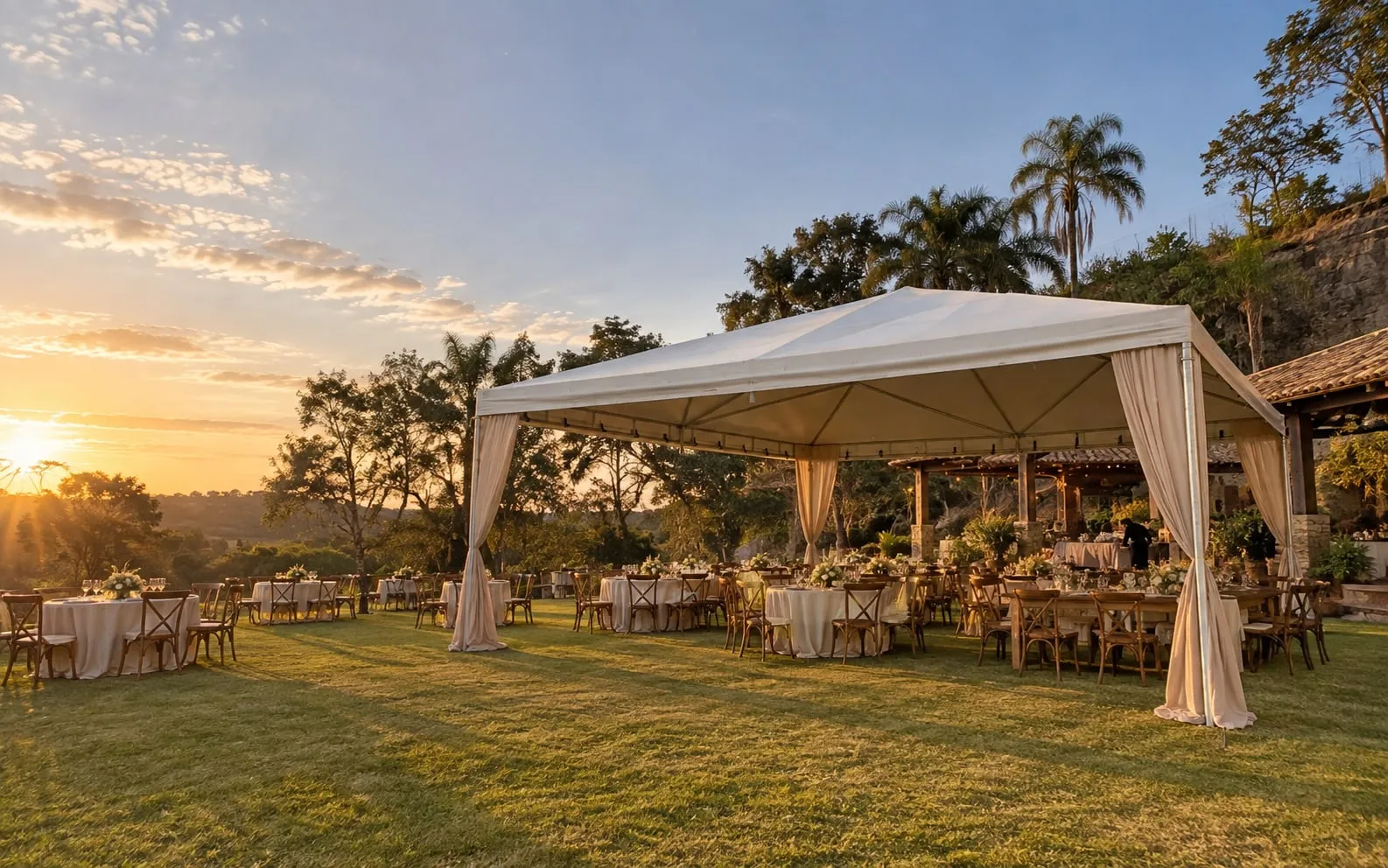Tenda com cortinas montada sobre gramado para recepção de casamento ao pôr do sol